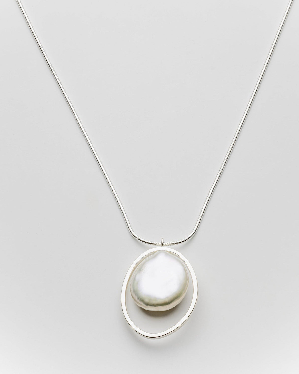 A silver necklace featuring a large, irregular white baroque pearl suspended inside a thin, open oval ring, suspended from a delicate silver chain against a light gray background.