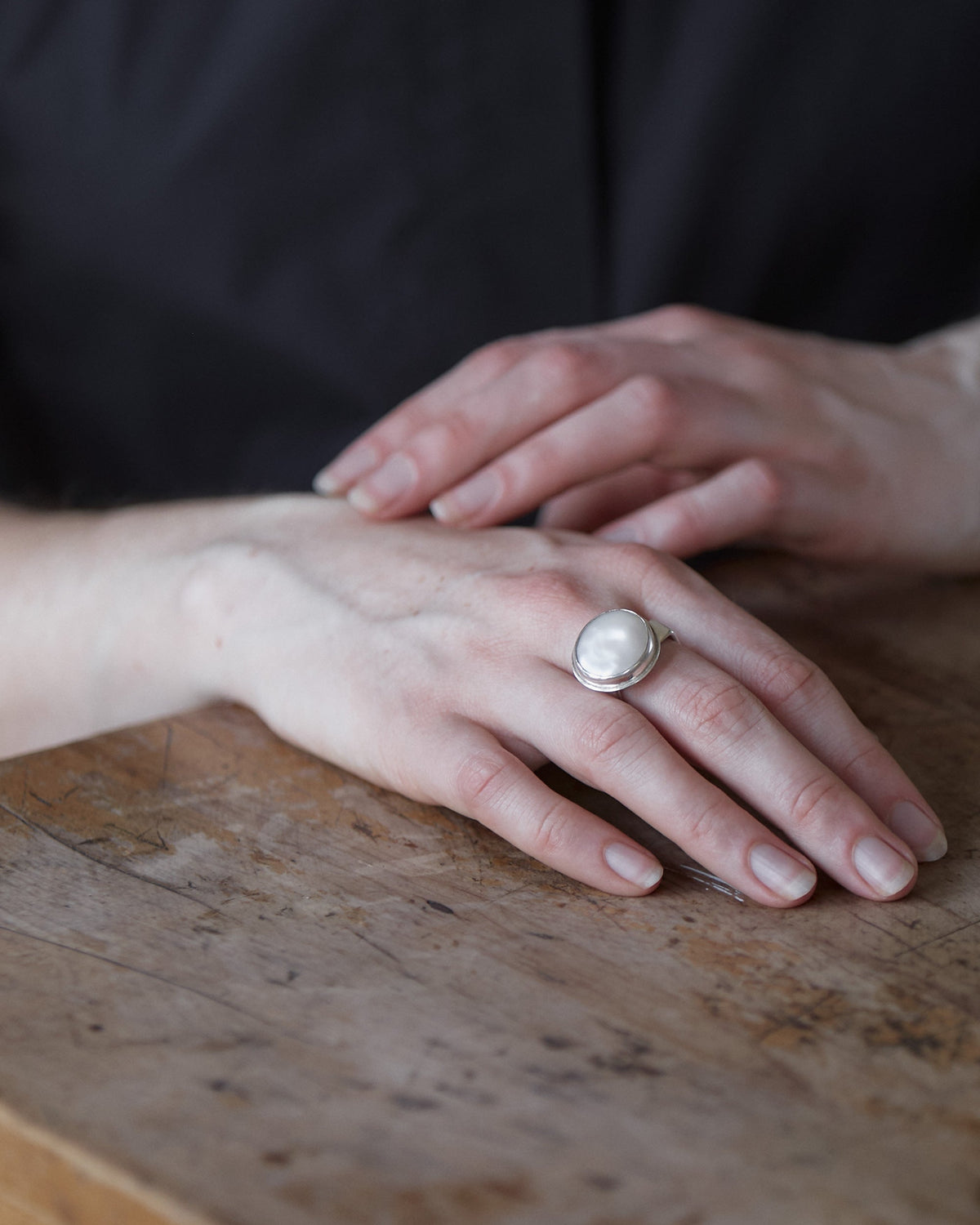 A person's hand wearing a sterling silver ring with a freshwater pearl, placed on a wooden surface.