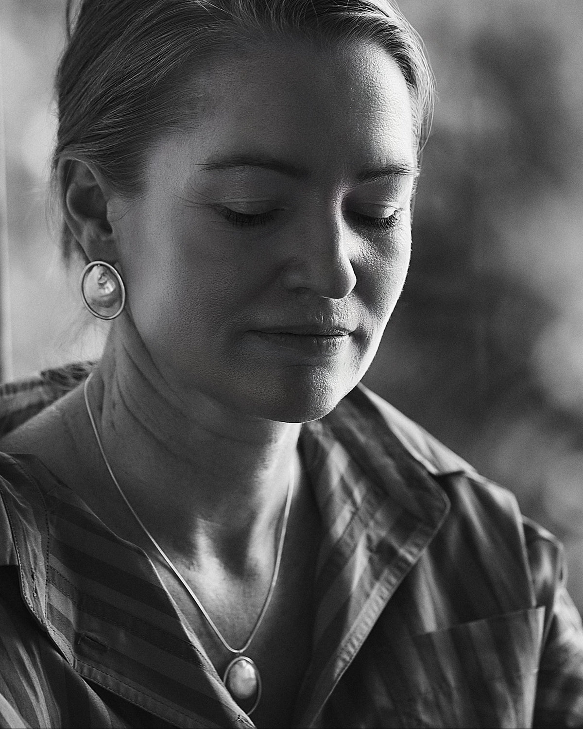 A black and white close-up portrait of a woman looking downward, wearing a striped collared shirt, a round pendant necklace, and a large circular earring.