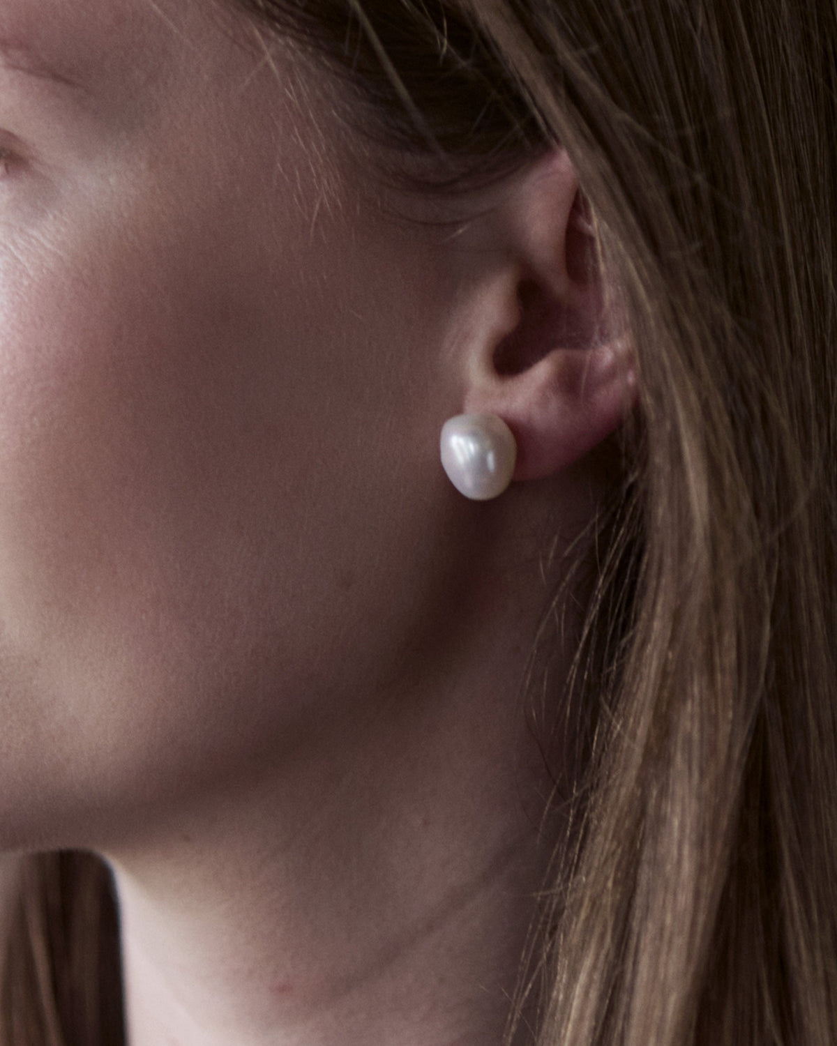 A close-up image of a woman's ear wearing a silver stud earring with a white baroque freshwater pearl.