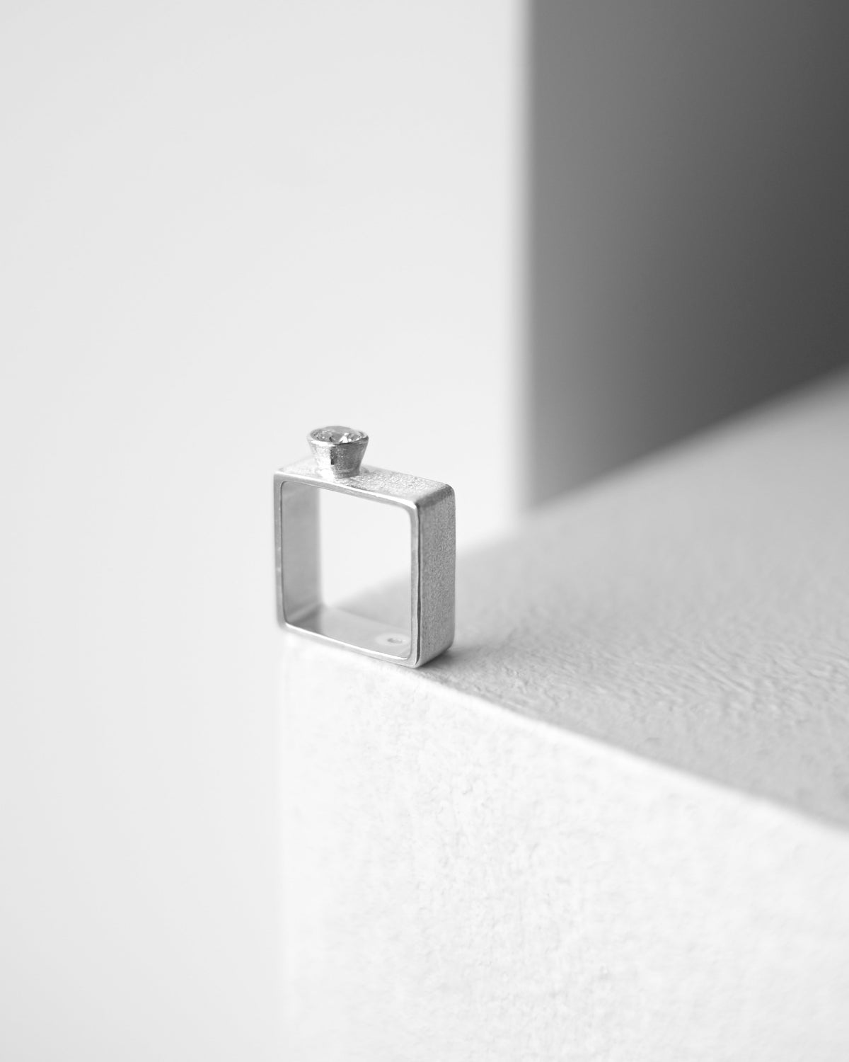 A minimalist silver square ring with a gem, resting on a white textured surface in soft, geometric lighting.