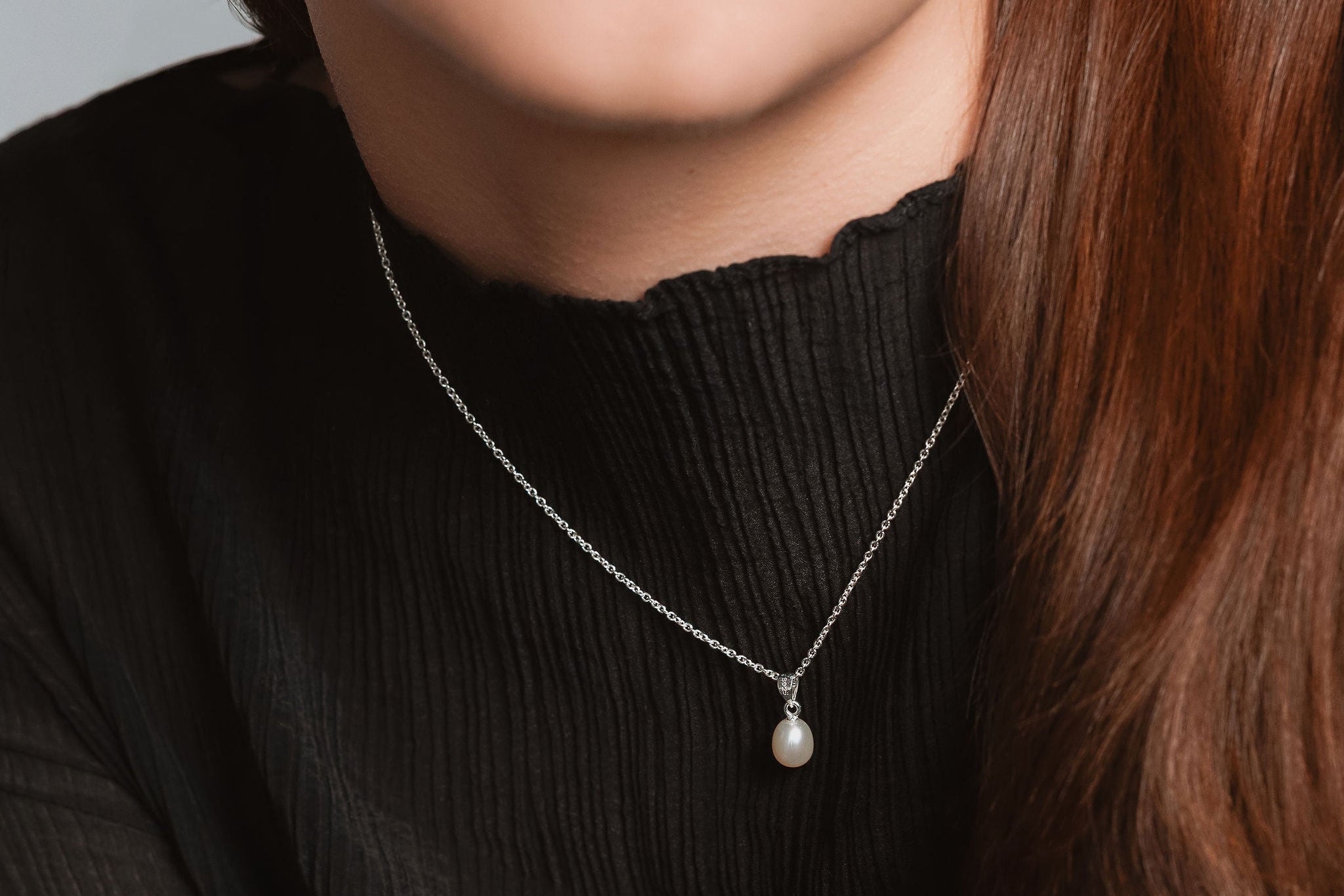 Close-up of a woman wearing a black sweater and a necklace with a pearl pendant against a neutral background.