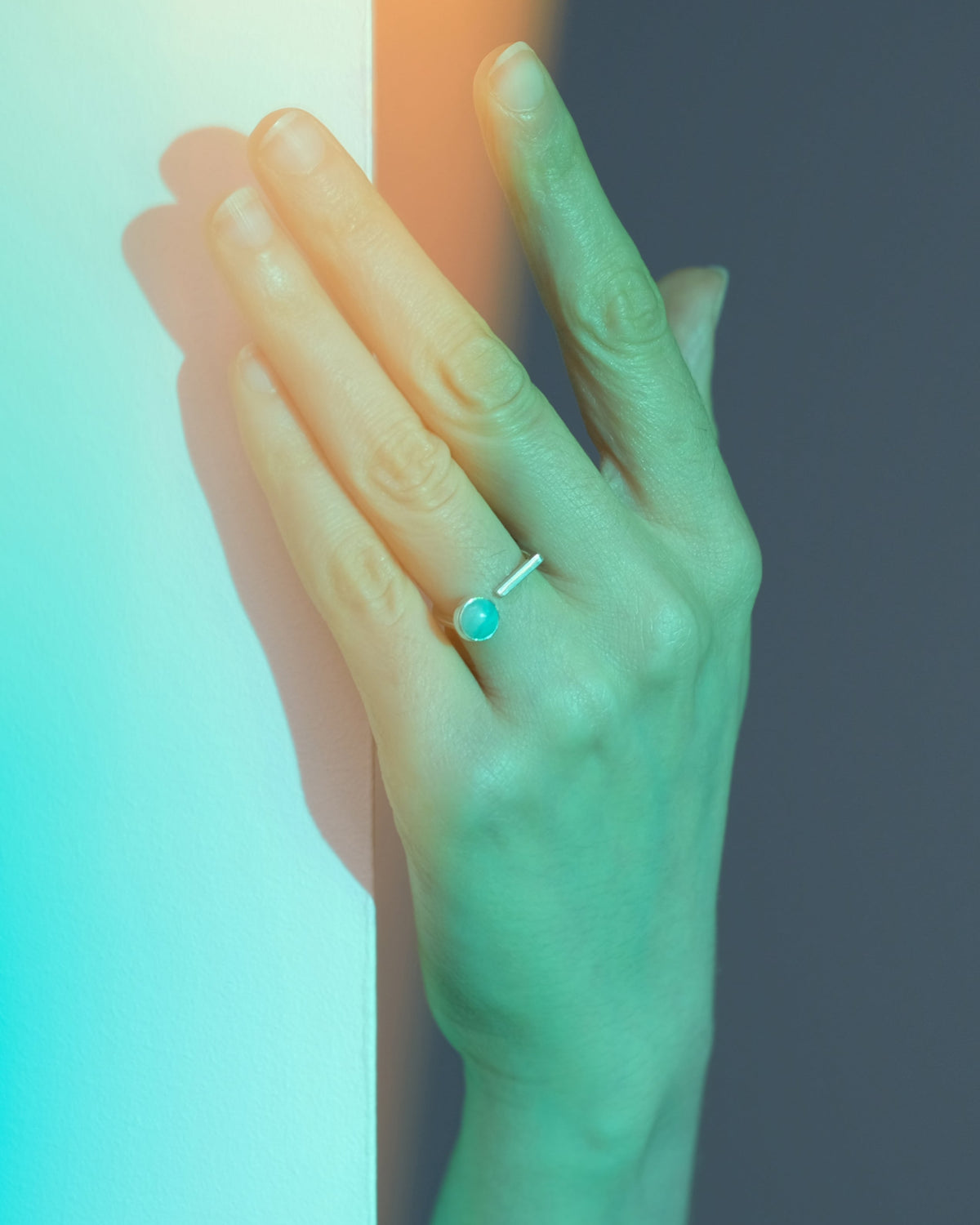 A close-up of a hand wearing a silver ring with a round amazonite gemstone, set against a split background of teal and dark gray with warm light reflections.