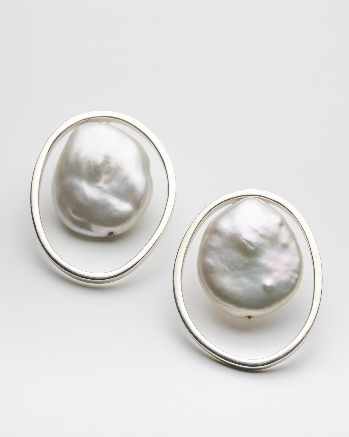A pair of silver-toned statement earrings featuring large, irregular baroque pearls set within open oval frames, photographed on a clean light gray background.