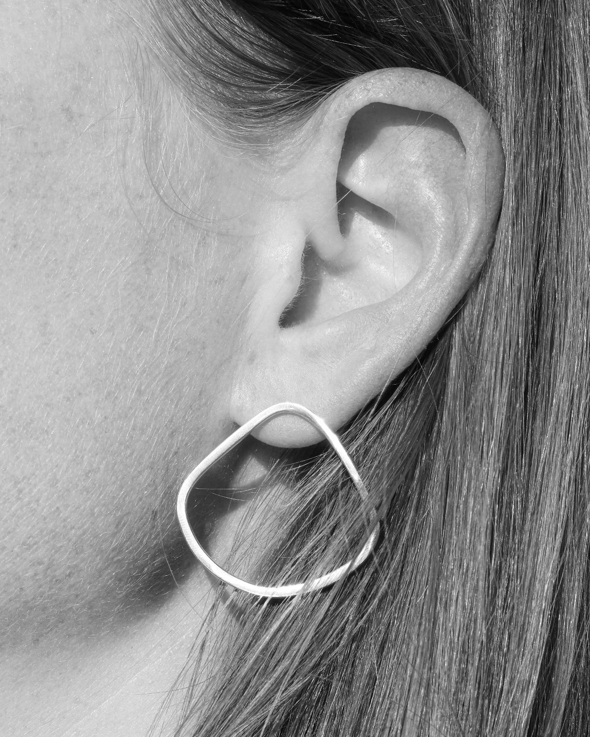 A close-up black and white photo of a person's ear wearing a thin triangular hoop earring.