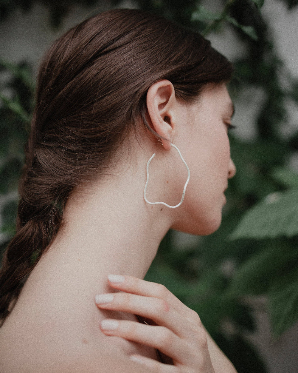 A woman in profile wearing a single large, organic-shaped silver hoop earring, with her hand resting on her shoulder against a soft-focus background of green leaves.