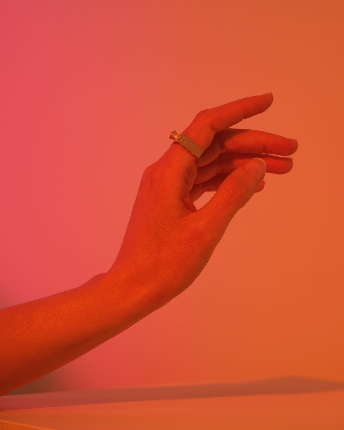 A close-up of a hand wearing a square silver band ring, set against a warm orange and pink gradient background.
