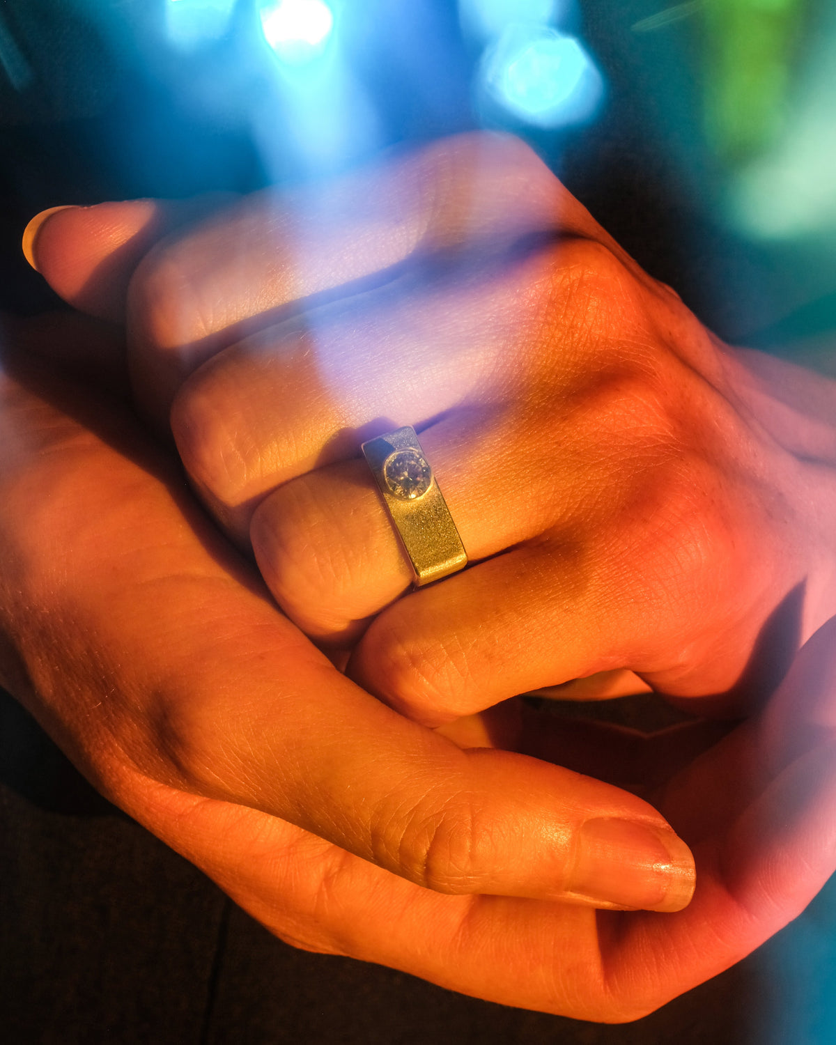 A close-up lifestyle shot of two hands holding each other, with a silver square band ring featuring a central clear gemstone worn on one finger. The image is illuminated by dramatic blue and orange lighting with soft bokeh in the background.